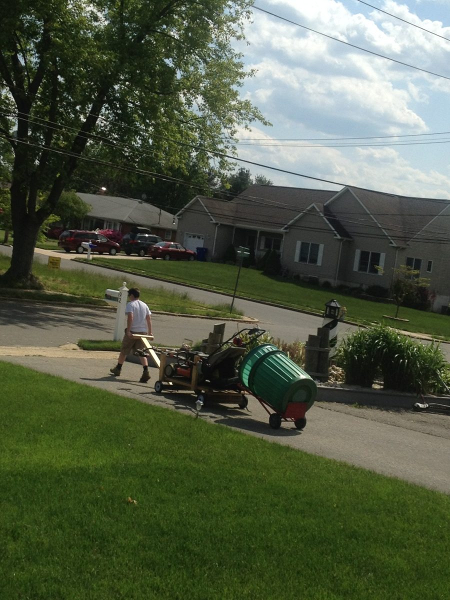 Young Logan pulling a homemade cart loaded with a push mower and trash can down the street